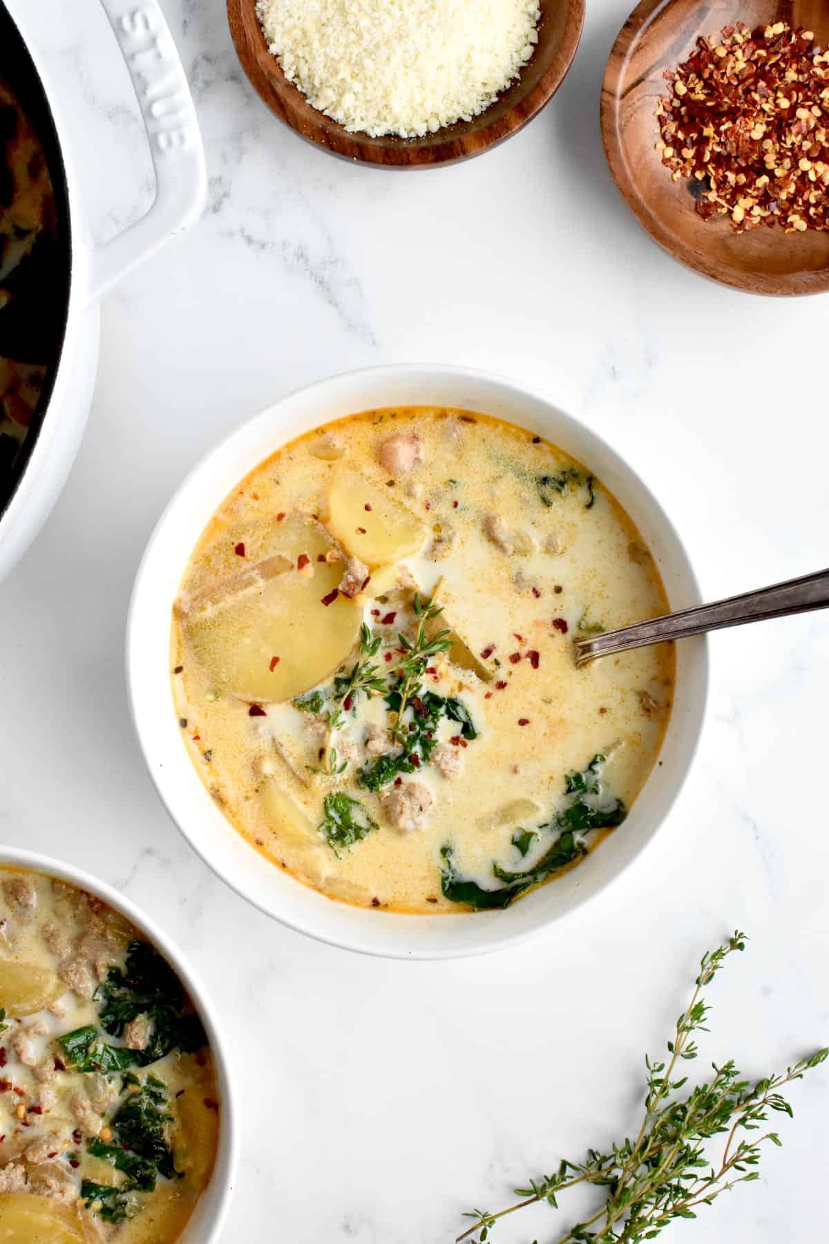 A bowl of zuppa toscana with a spoon in it, with another bowl of zuppa and pot to the side.