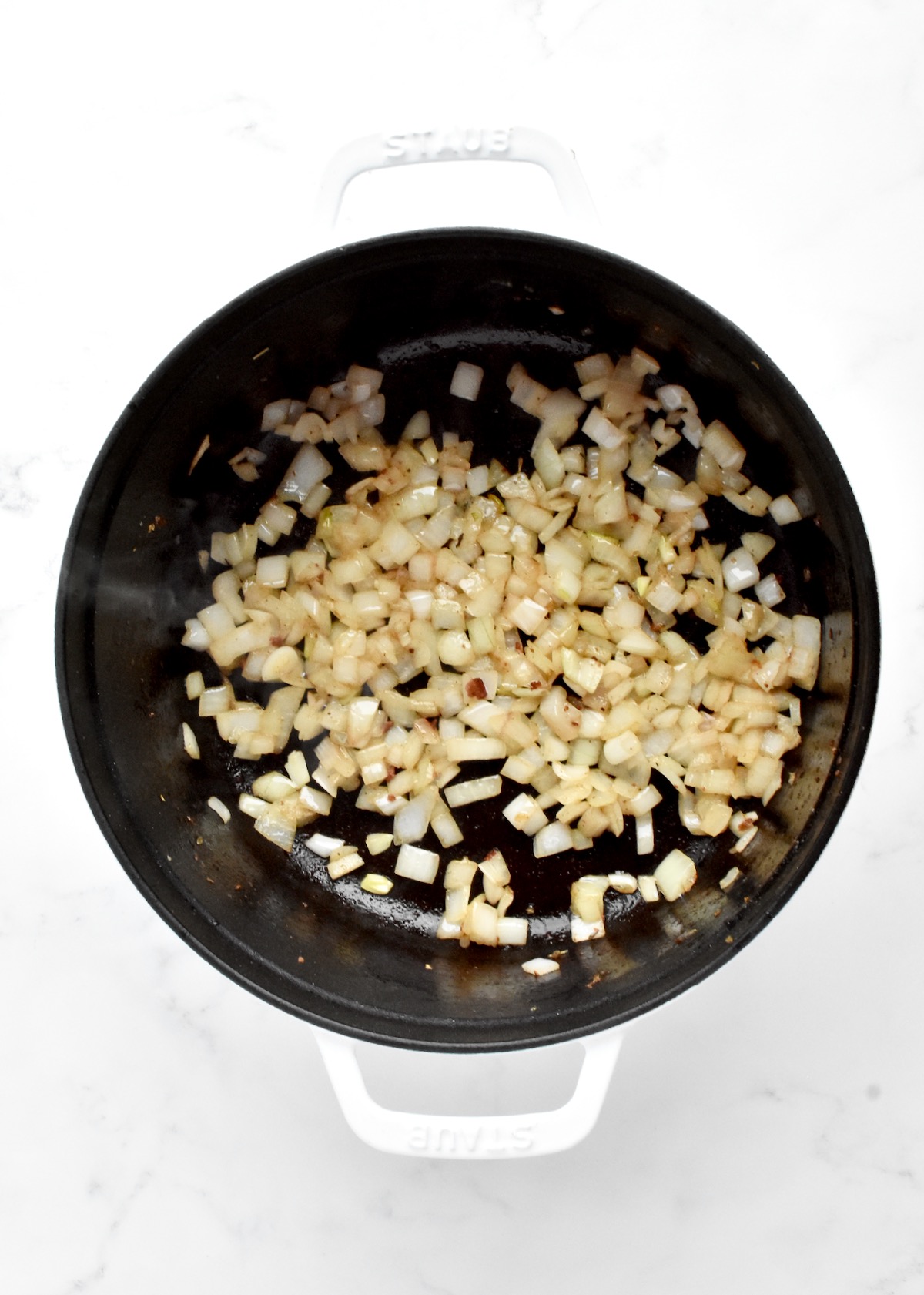 A white dutch oven on a marble background. The pot contains diced onions browning in bacon grease.