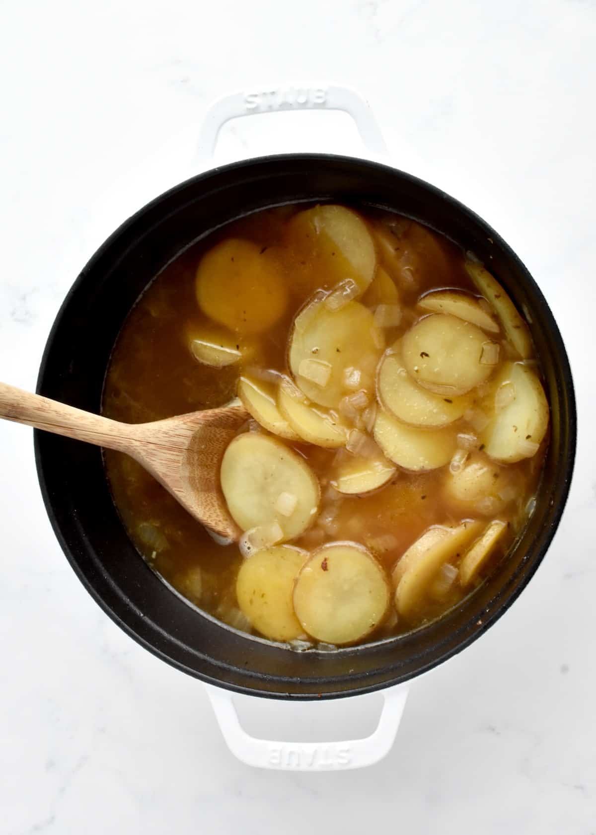 A white dutch oven on a marble background. The pot contains sliced potatoes in broth, stirred by a wooden spoon.