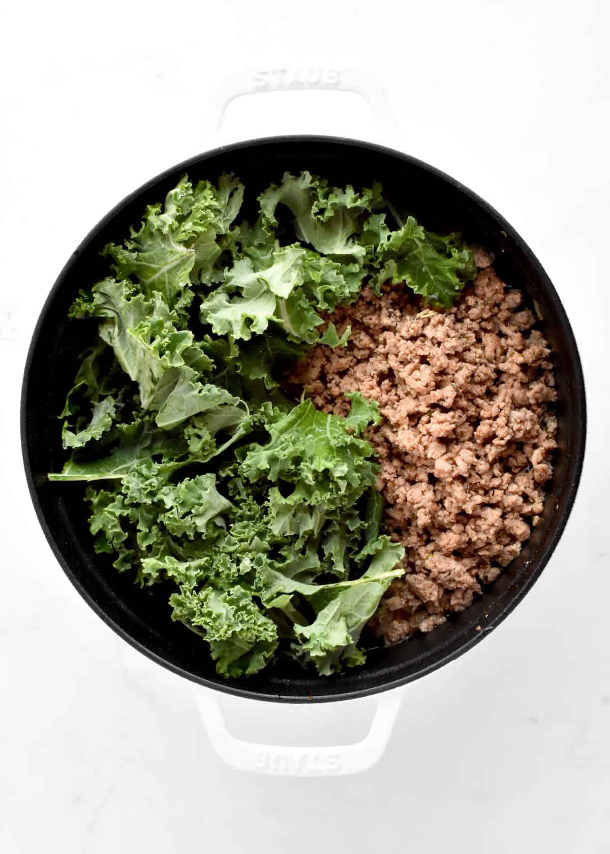 A white dutch oven on a marble background. The pot contains browned ground turkey and kale.