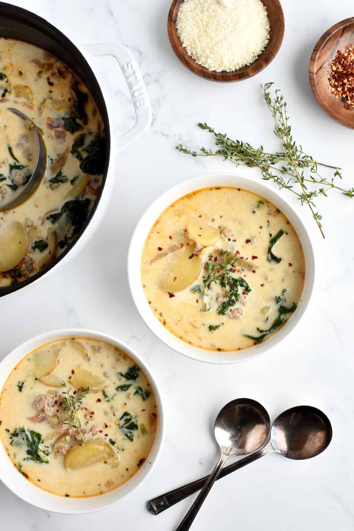 Two white bowls with healthy zuppa toscana, with a white pot of zuppa toscana and two garnish bowls to the side.