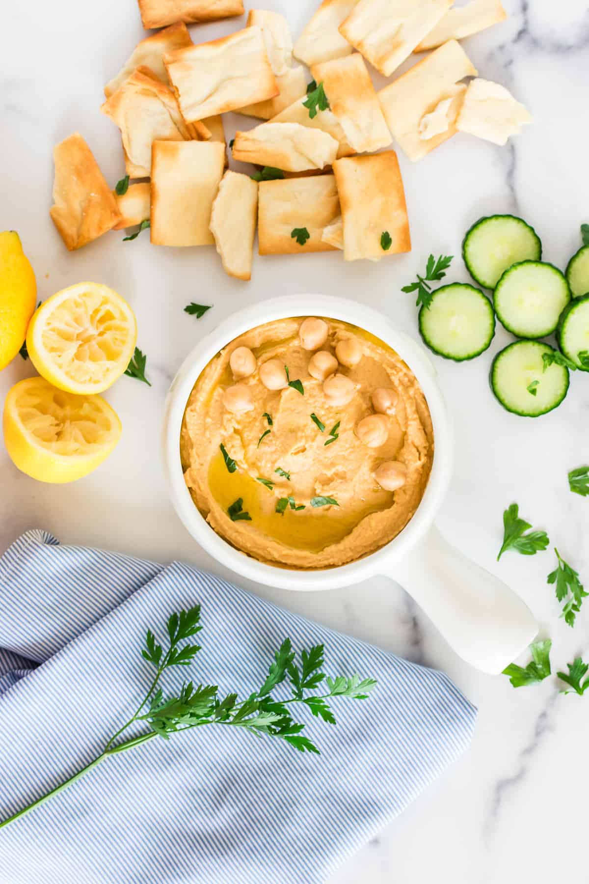 A small bowl of hummus with pita chips, cucumbers and lemons on the side, garnished with parsley.