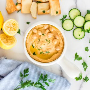 A small bowl of hummus with pita chips, cucumbers and lemons on the side, garnished with parsley.