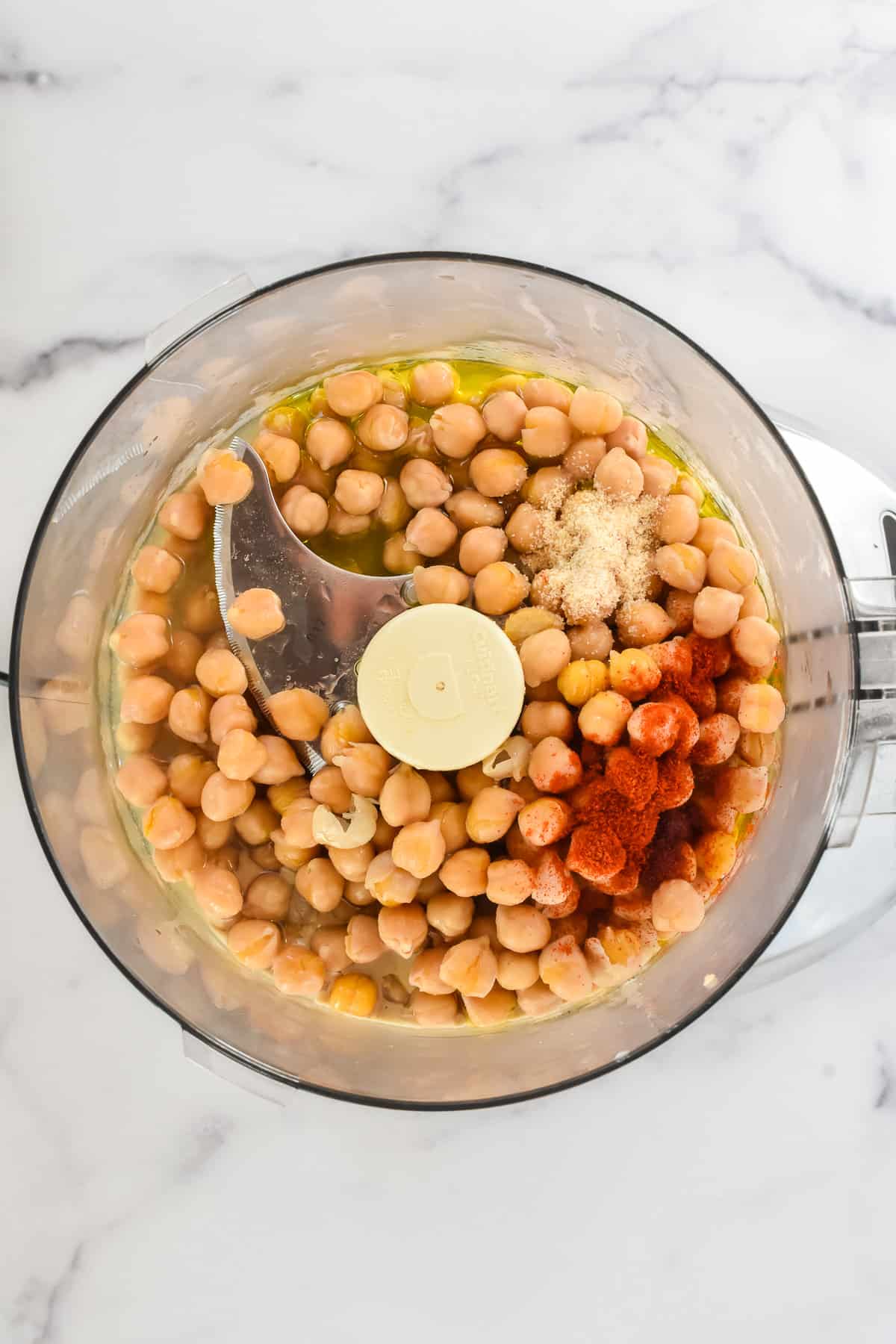 A close up view of the inside of a food process, with whole chickpeas and other ingredients for hummus in it, not yet blended.