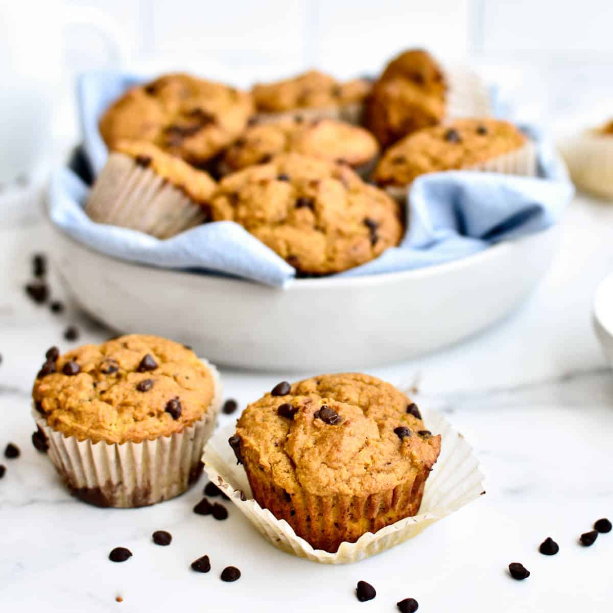 Two pumpkin chocolate chip muffins in the foreground, with a bowl of muffins behind them. One muffin is unwrapped from its wrapper and chocolate chips are scattered around.