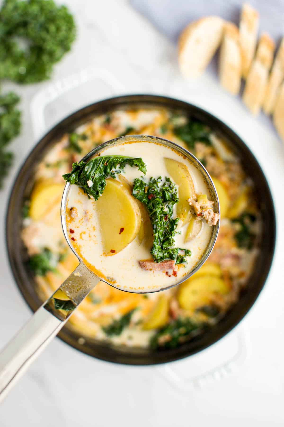 A close up of a ladle of soup over a white pot of zuppa toscana on a marble background.