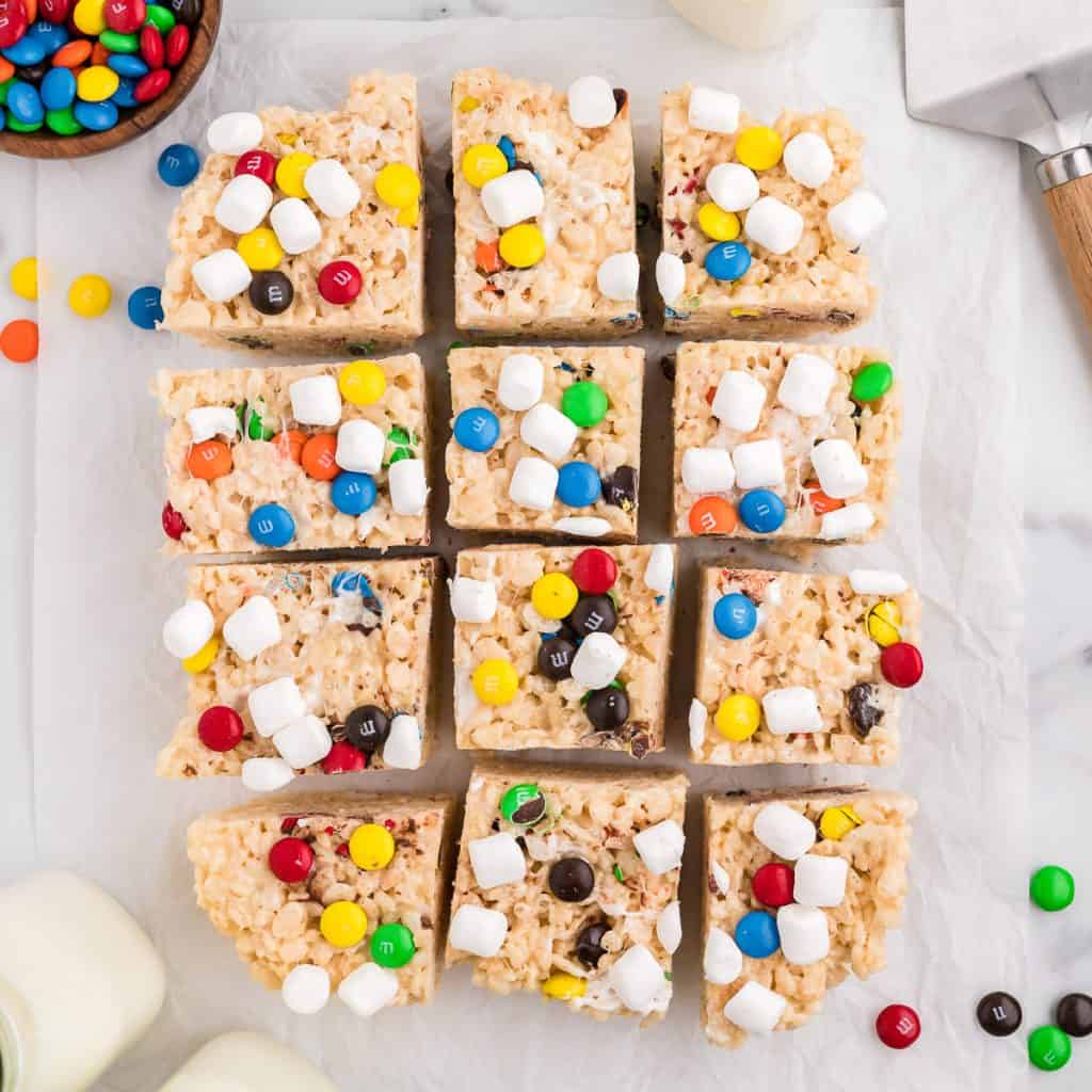 An overhead view of a 12 M&M rice krispie treats on a piece of parchment paper, with m&m candies scattered around and bottles of milk to the side.