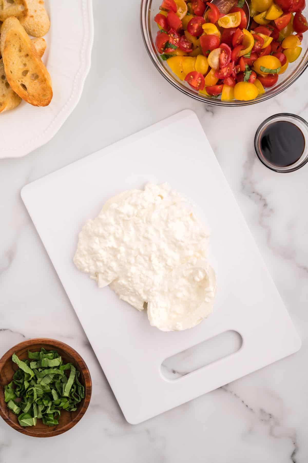 A white cutting board with a cut open burrata cheese ball, with the creamy center pouring out.