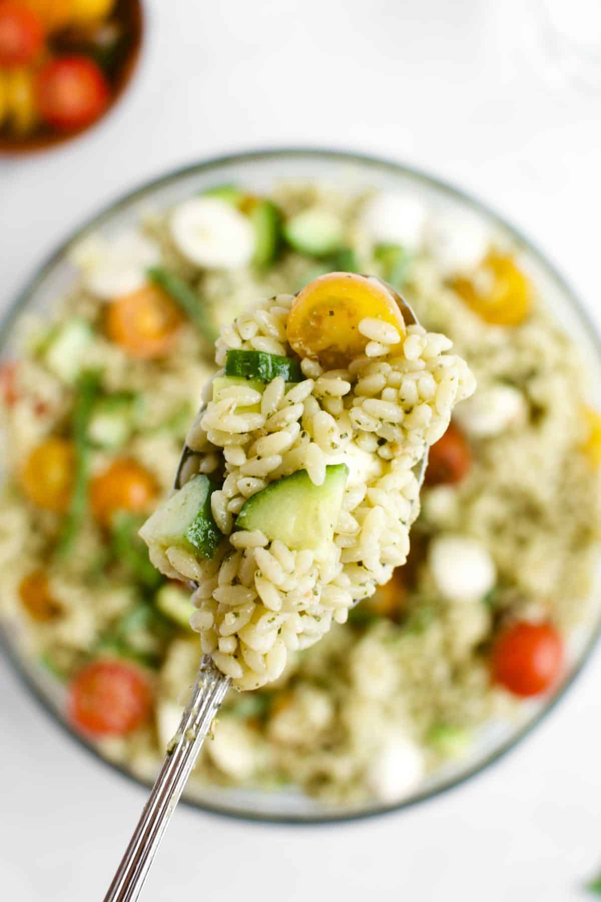 A spoon holding a scoop of orzo pesto pasta salad, with a bowl of pasta salad in the background.