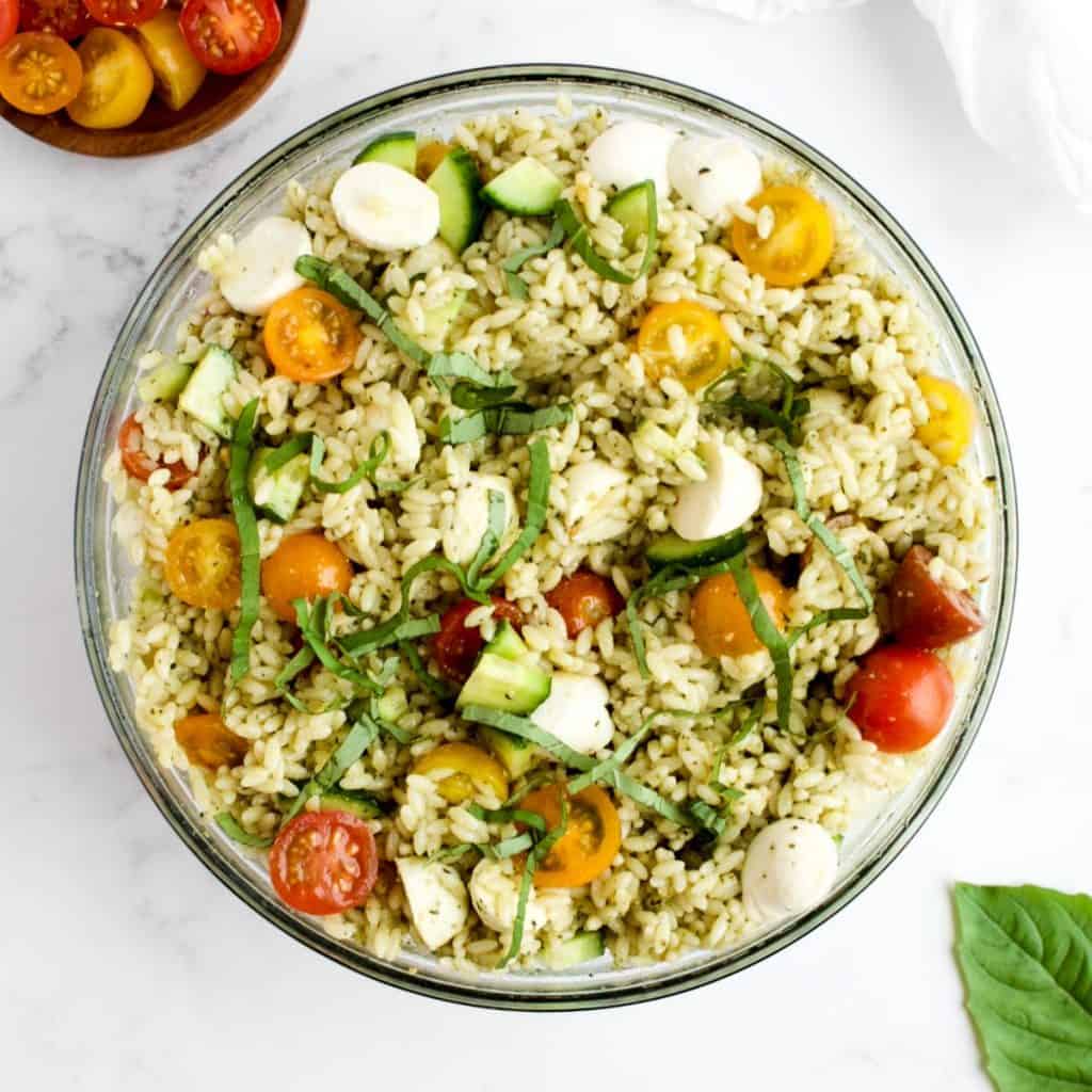 Orzo pesto pasta salad in a glass bowl on a marble background. Leaves of basil and a dish of cherry tomatoes are to the side.