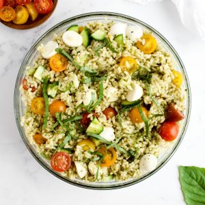 Orzo pesto pasta salad in a glass bowl on a marble background. Leaves of basil and a dish of cherry tomatoes are to the side.