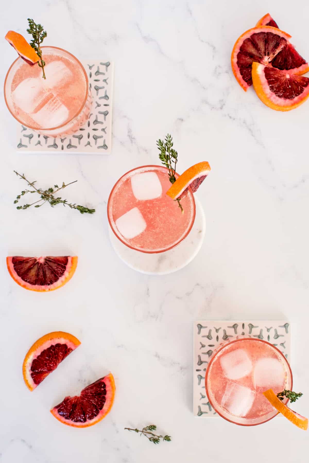 A top down view of three old fashioned cocktails on a marble background, with cut blood oranges and sprigs of thyme as garnish.