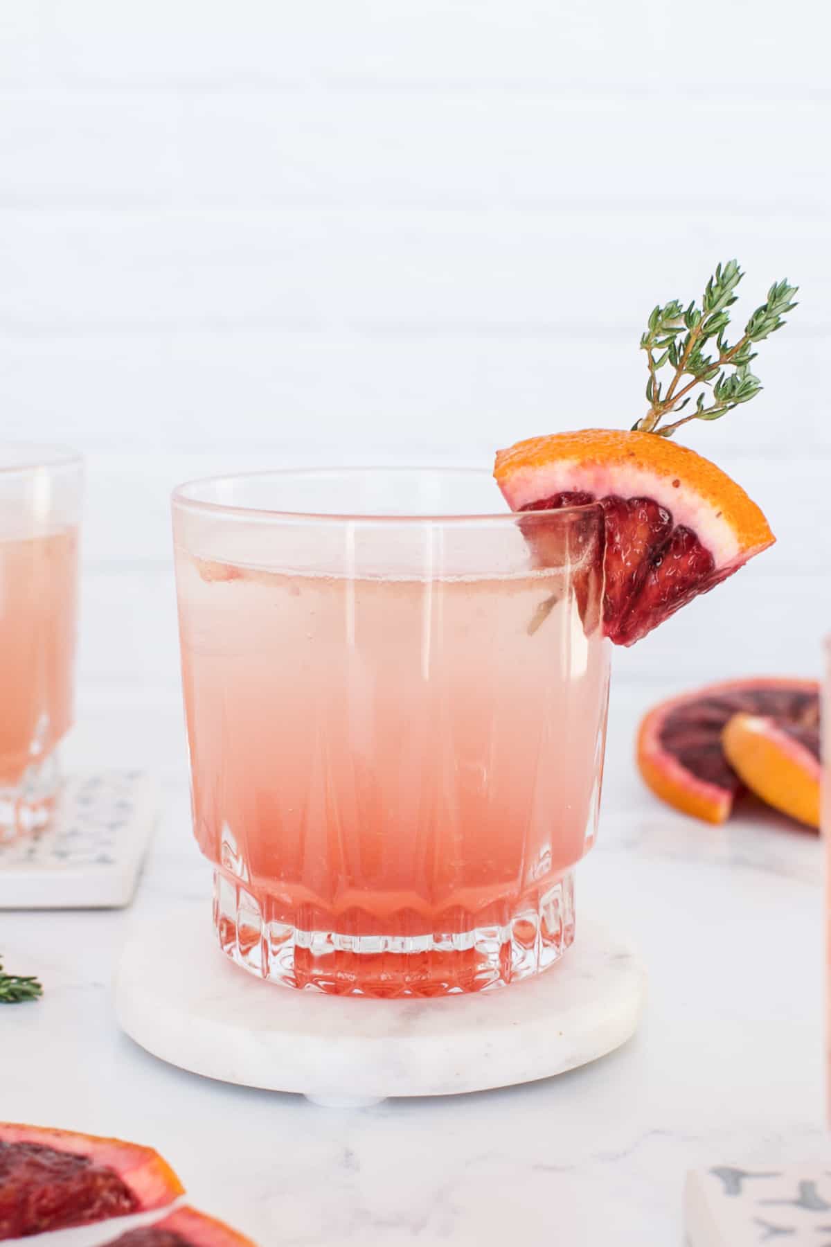 A rocks glass containing a pink blood orange old fashioned cocktail, garnished with a wedge of blood orange.