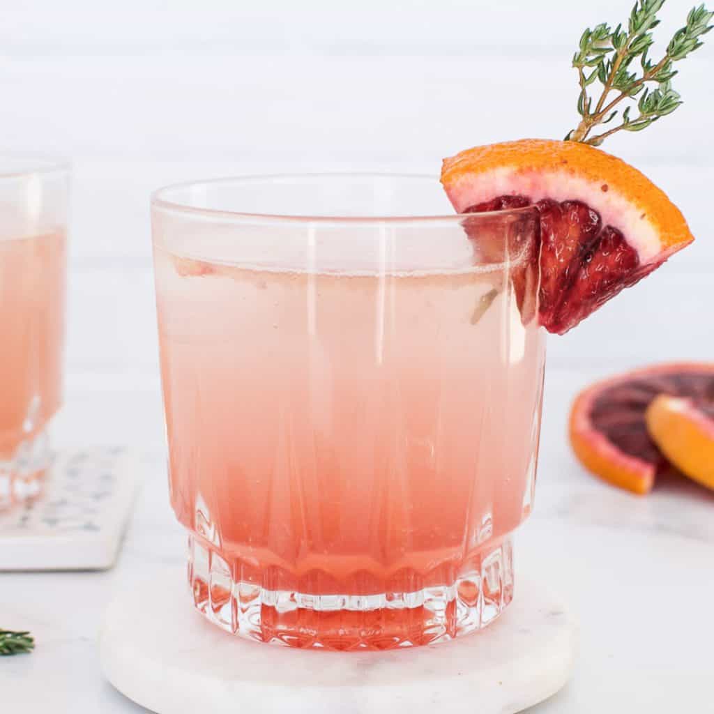 A rocks glass containing a pink blood orange old fashioned cocktail, garnished with a wedge of blood orange.