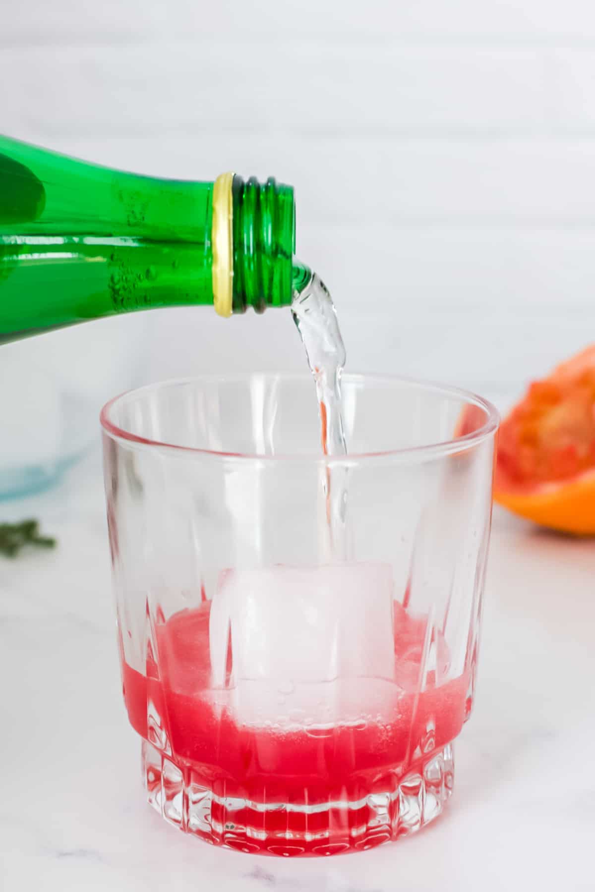 A rocks glass on a marble background with sparkling water being poured into a pink cocktail with a large ice cube.