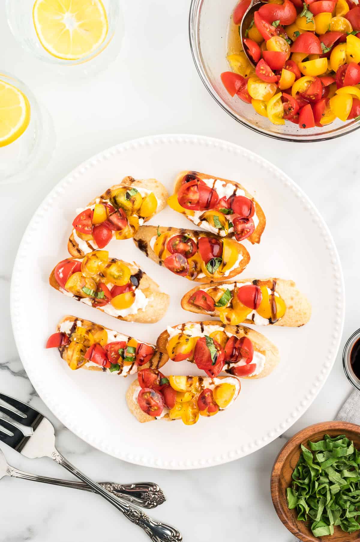 A round white platter with 8 bruschetta toasts on a marble background. Cherry tomato mixture and fresh basil sits to the side.