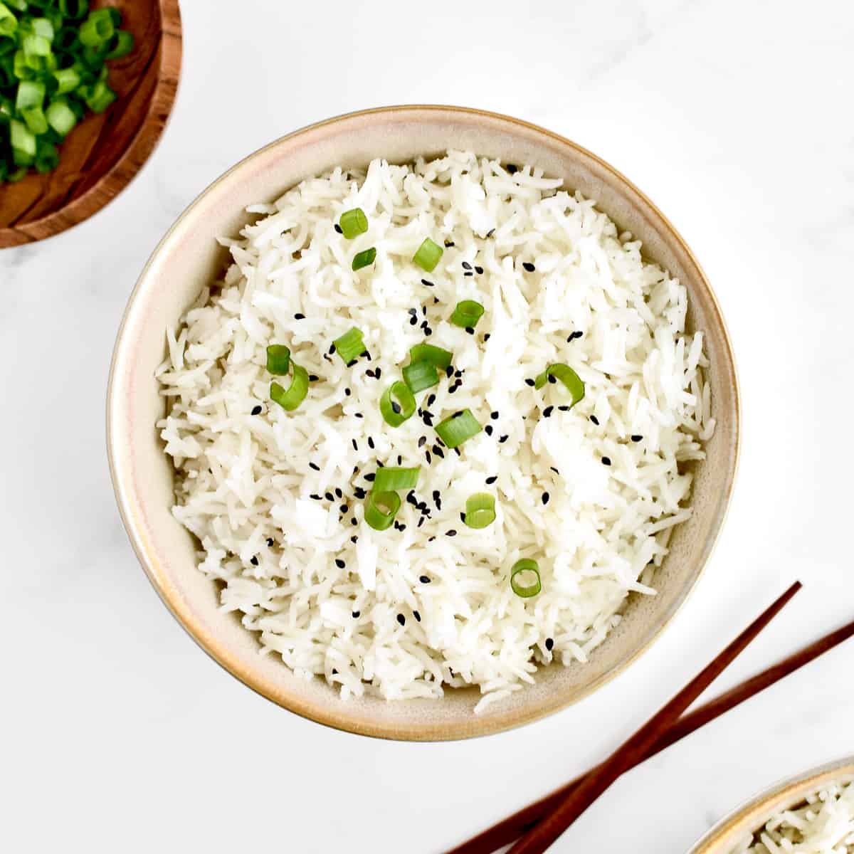 A bowl of coconut basmati rice, garnished with sesame seeds and green onions, on a marble background. Chopsticks sit to the side of the bowl.