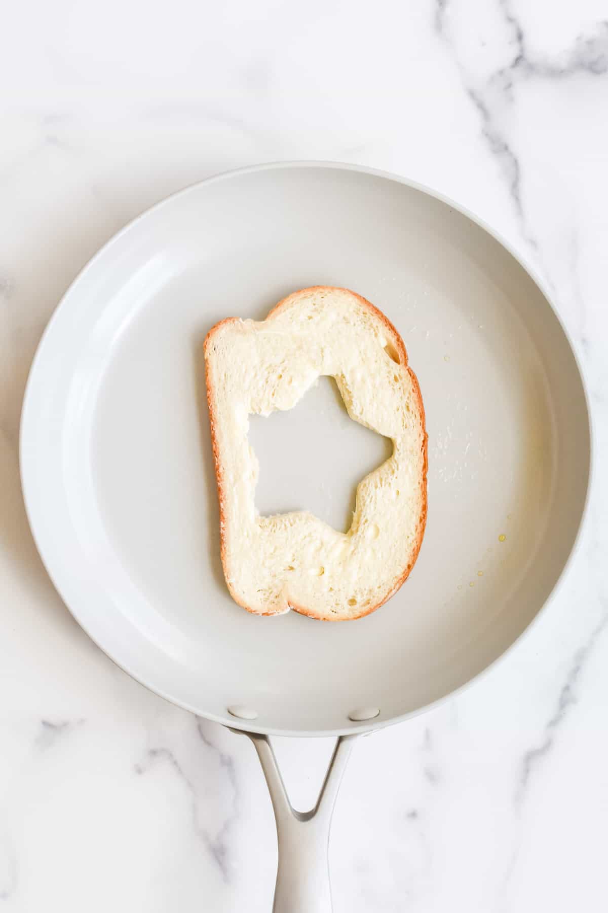 A grey skillet on a marble background. The skillet contains a slice of buttered bread with a star cut from the middle.