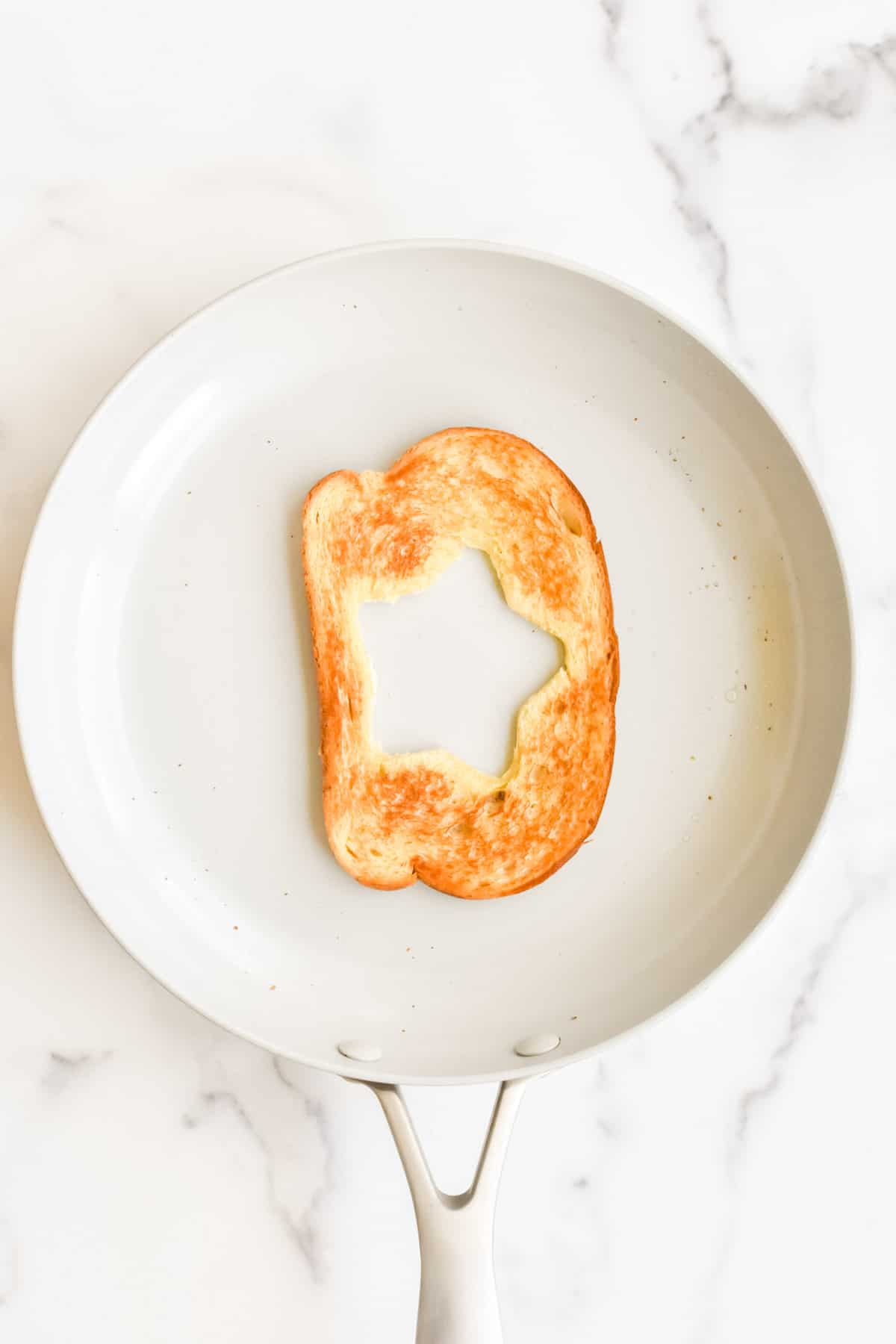 A grey skillet on a marble background. The skillet contains a slice of buttered bread with a star cut from the middle with the top side toasted.
