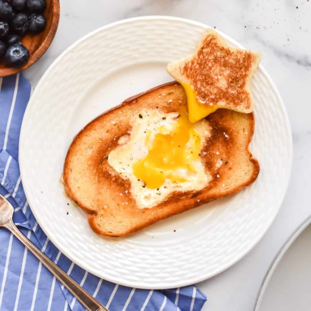 A white plate on a marble background. The plate contains a piece of toast with a cooked egg in a star shaped hole. A toasted star shaped bread is to the side, dipped into the egg yolk.