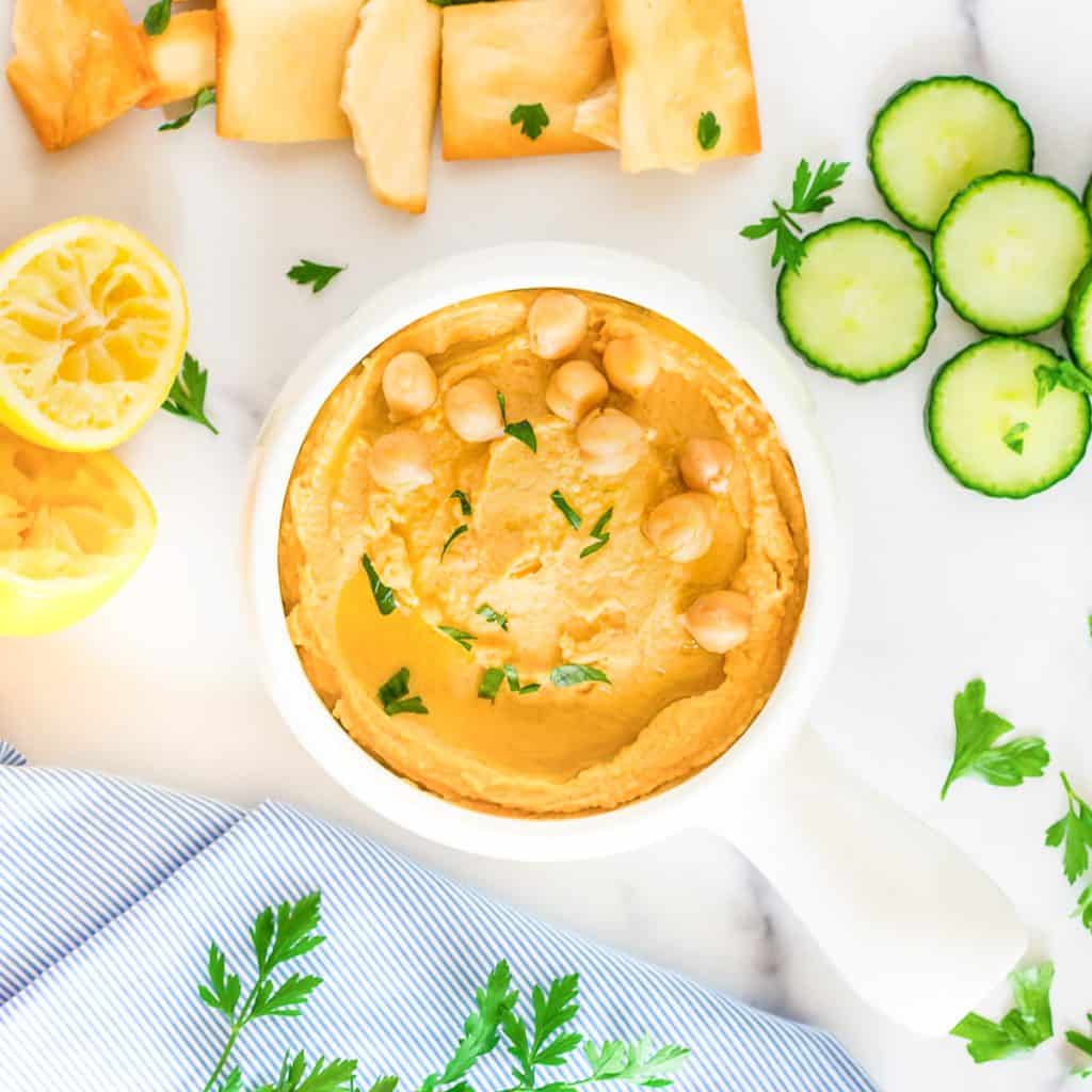 A white dish of hummus on a marble background, with parsley, lemon, cucumber slices and pita chips at the side.