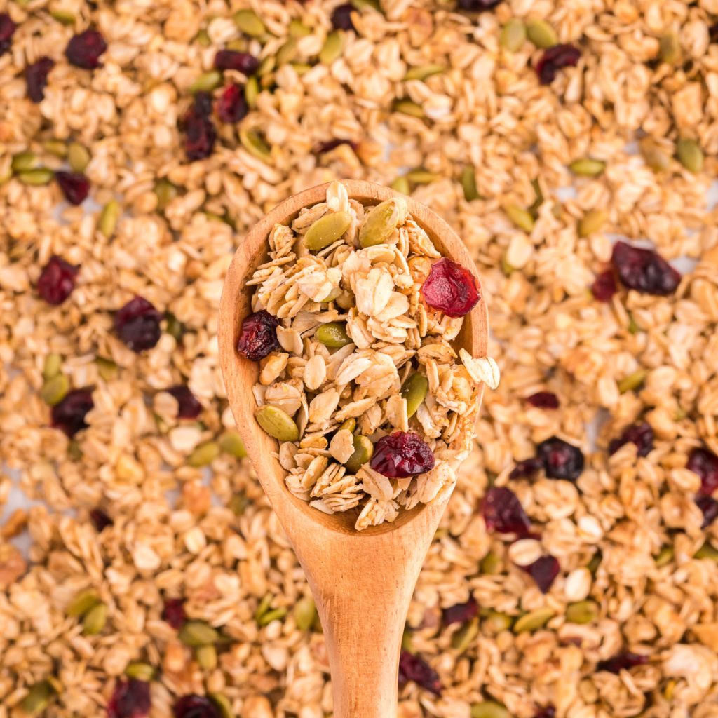 A close up of a wooden spoon with a scoop of maple walnut granola dotted with cranberries and pepitas.