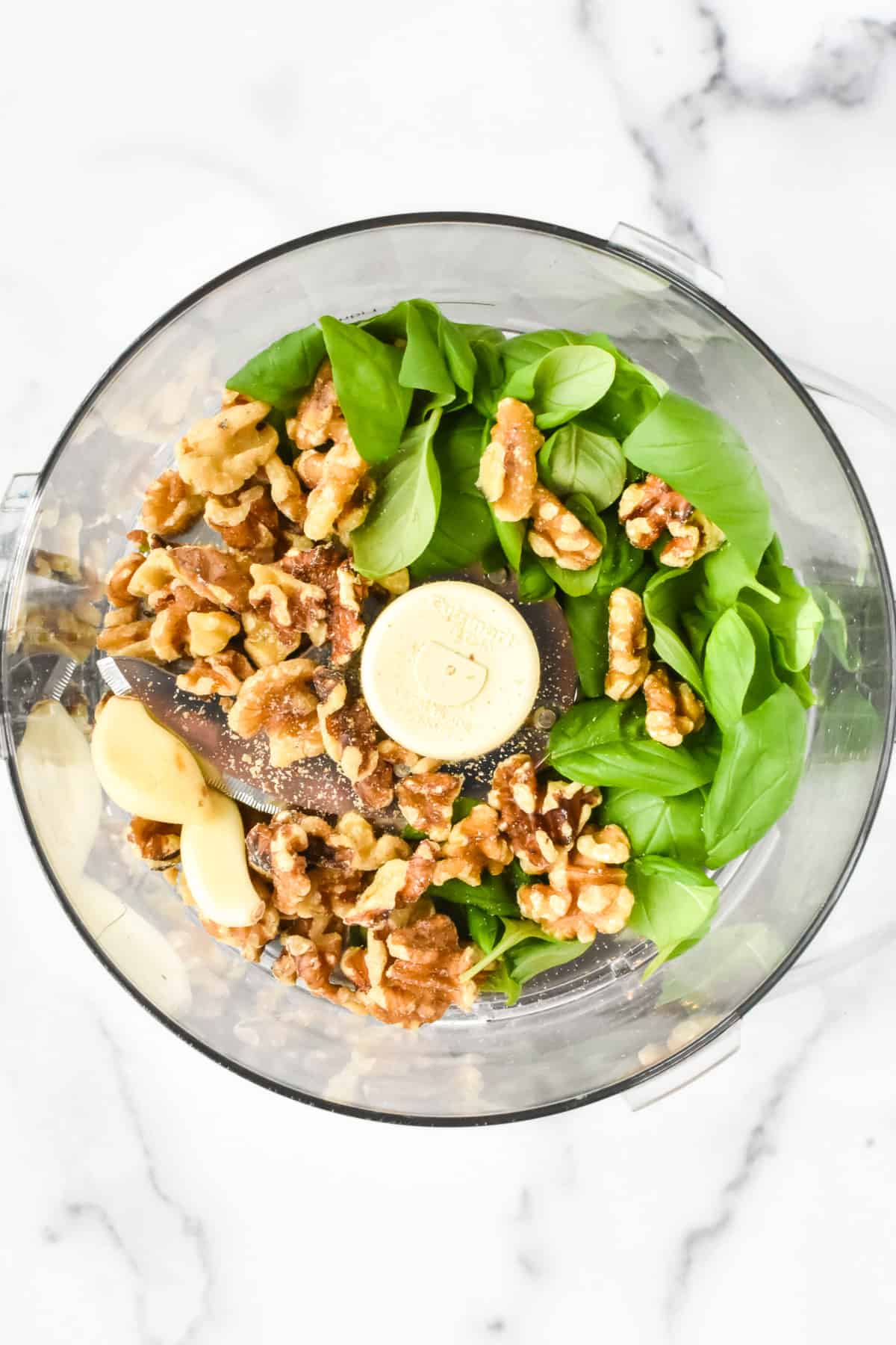 The inside of a food processor containing basil, walnuts and garlic on a marble background.