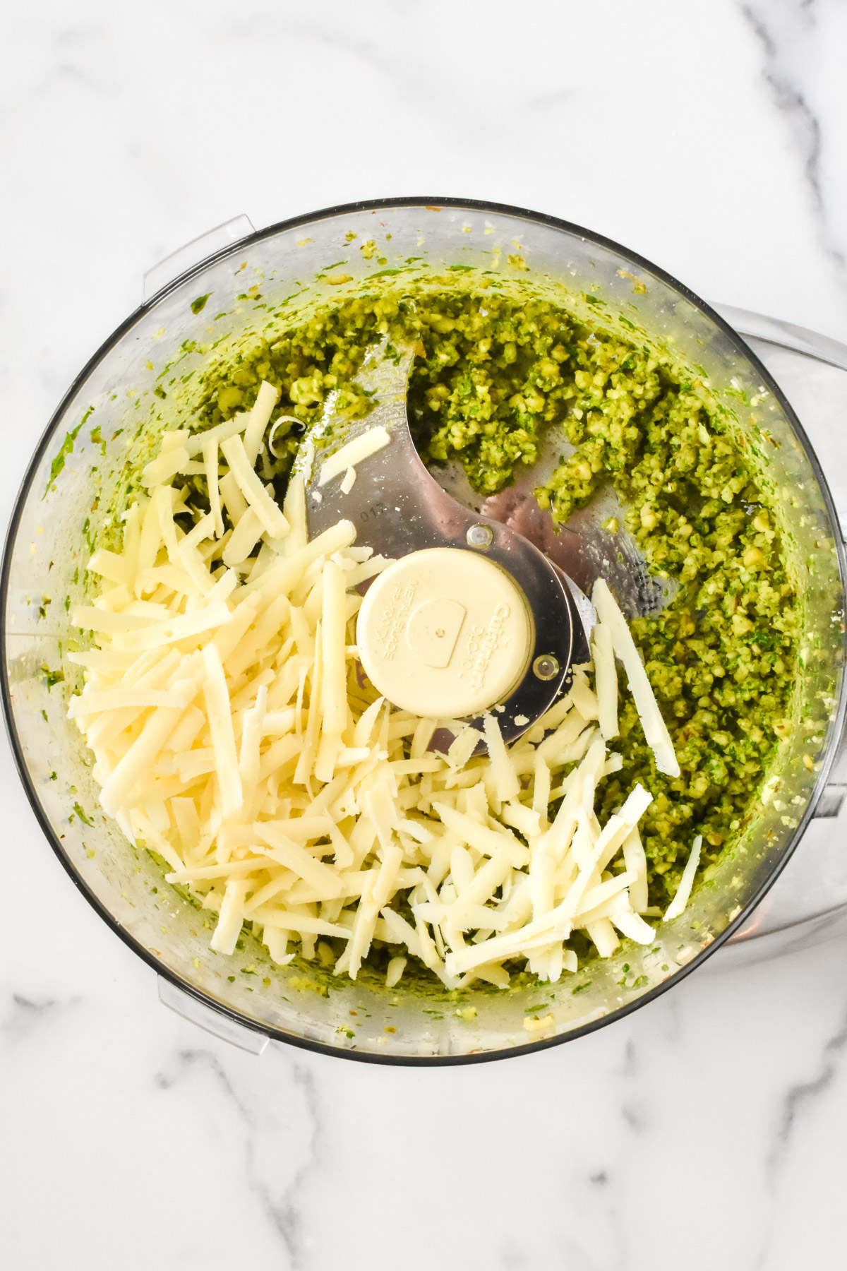 The inside of a food processor containing blended walnuts and bail, topped with shredded parmesan.