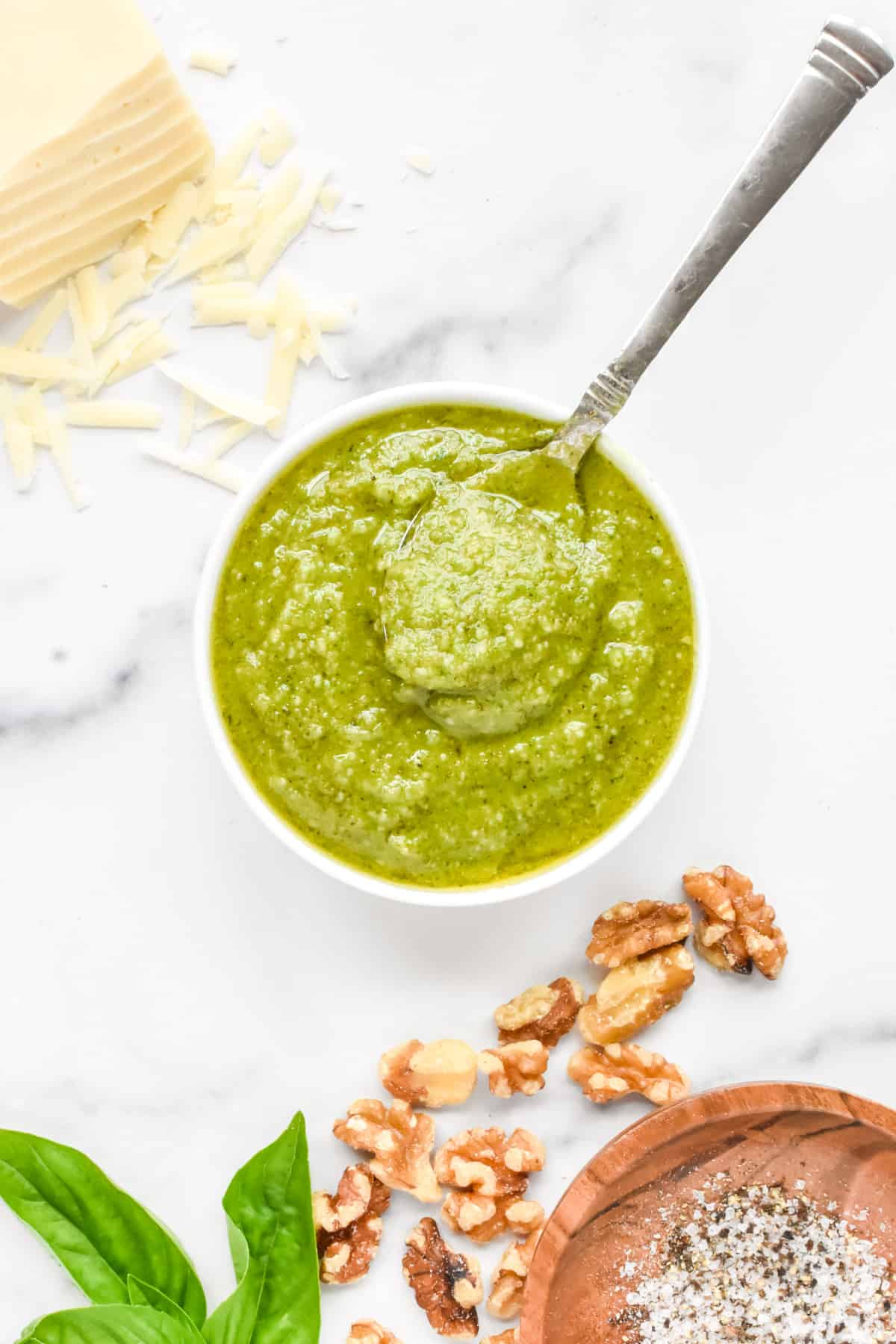 A small white bowl of basil pesto without pine nuts, with basil, walnuts and cheese to the side.