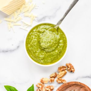 A small white bowl of basil pesto without pine nuts, with basil, walnuts and cheese to the side.