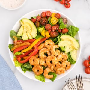 A white bowl with layers of romaine, shrimp, fajita veggies, cherry tomatoes and avocado.