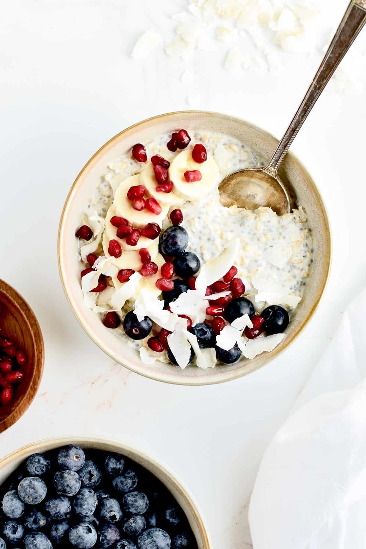 A grey bowl of vanilla overnight oats on a marble background, garnished with banana, berries, coconut and pomegranate seeds.