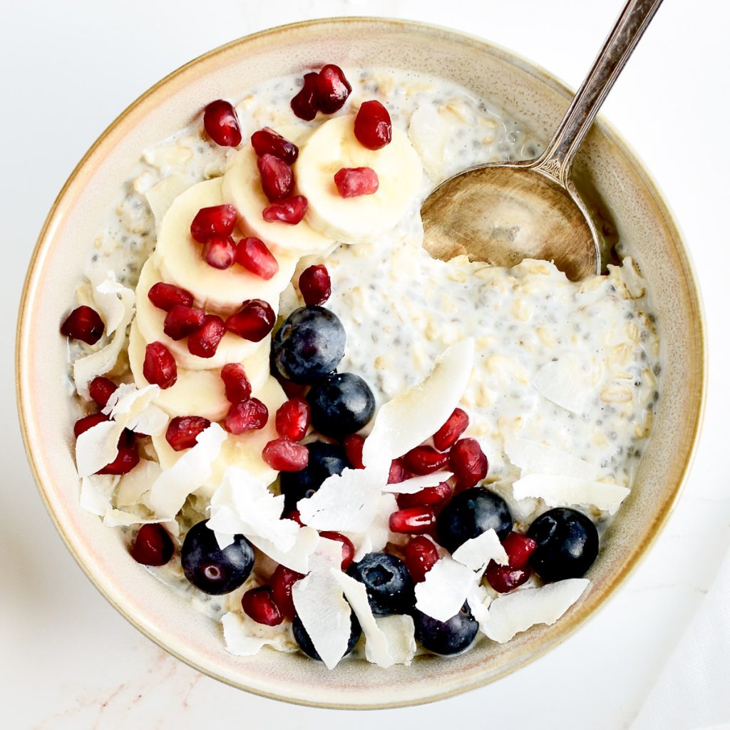 A grey bowl of vanilla overnight oats on a marble background, garnished with banana, berries, coconut and pomegranate seeds.