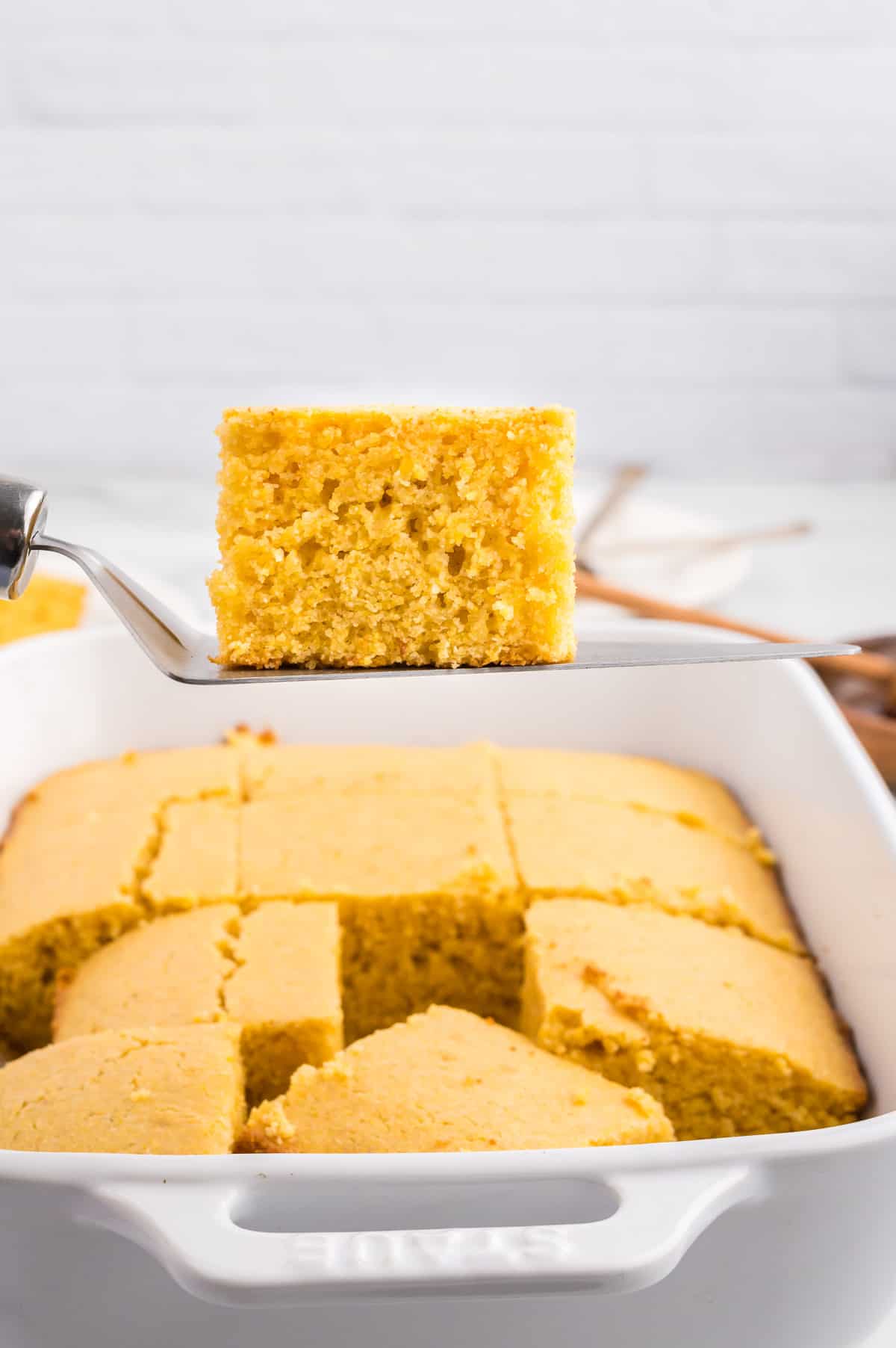 A slice of cornbread on a pie server, with a pan of cornbread beneath it.