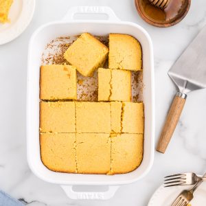 A pan of cornbread, with several slices removed, with plates of cornbread and a dish of honey to the side.