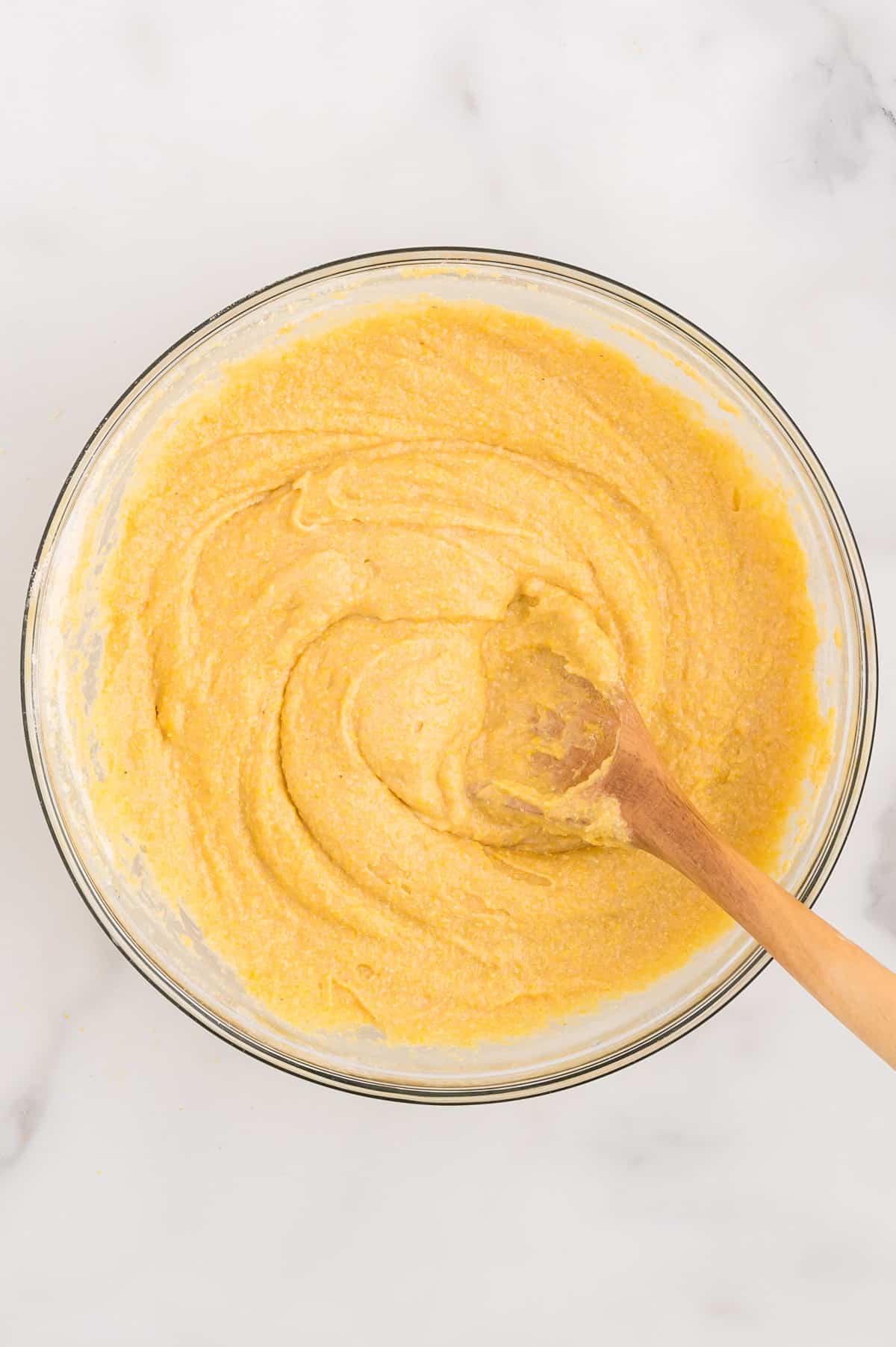 A glass bowl with cornbread batter being stirred by a wooden spoon.