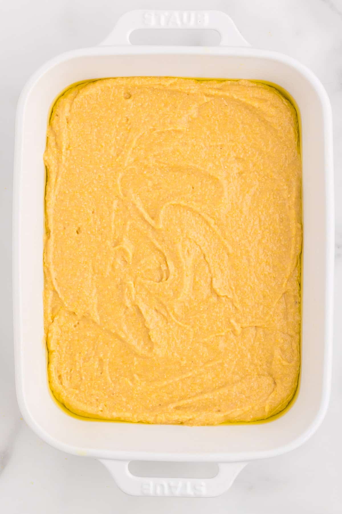 A white baking dish with unbaked whole grain cornbread on a marble background.