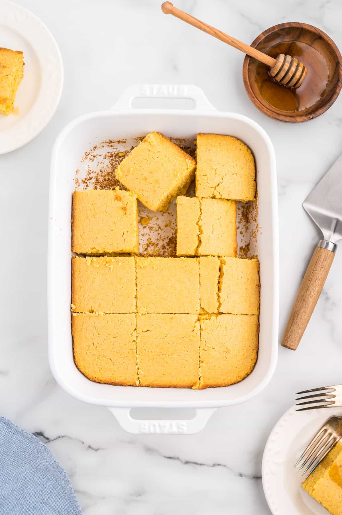 A pan of cornbread, with several slices removed, with plates of cornbread and a dish of honey to the side.