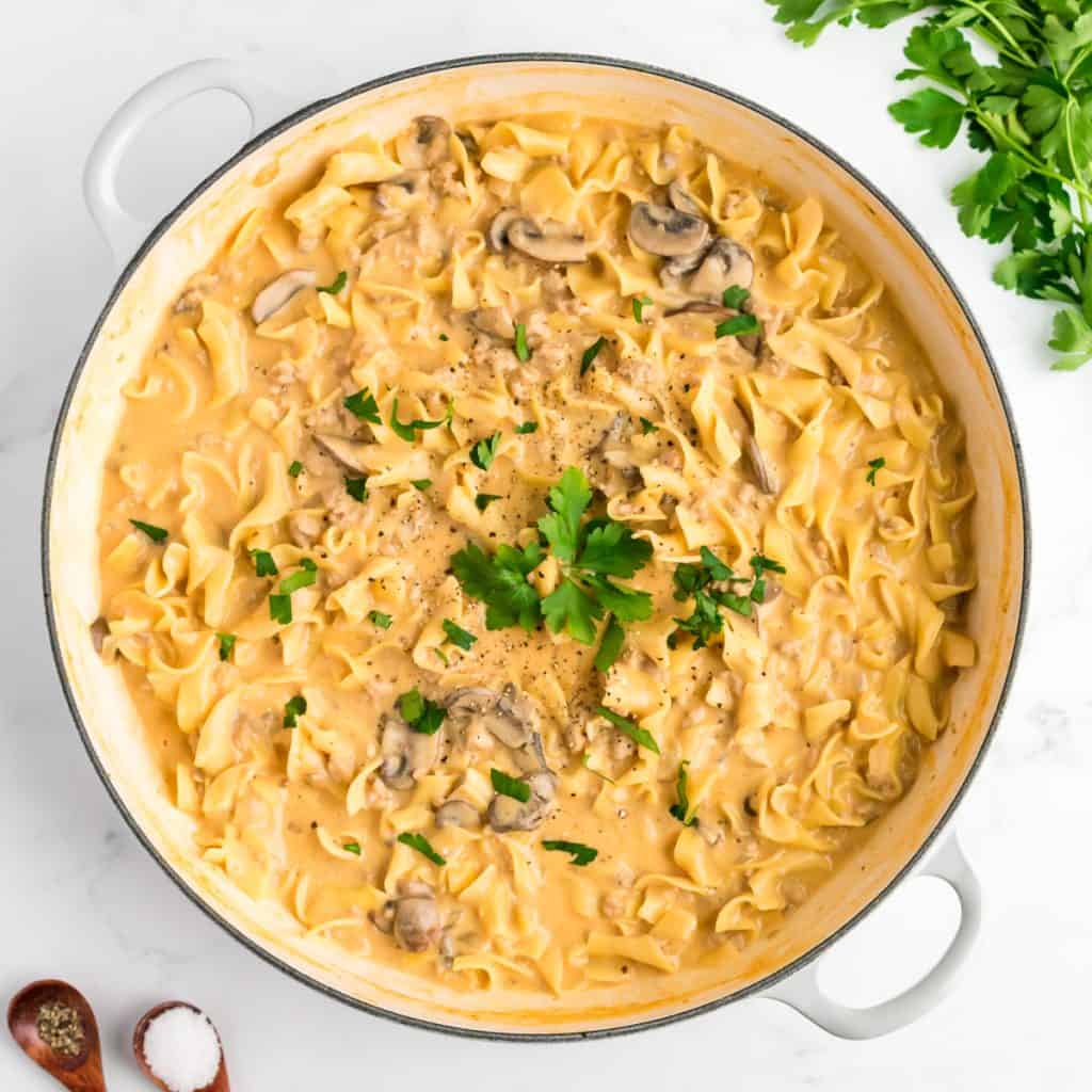 A white skillet of creamy ground chicken stroganoff on a marble background. Two small spoons of salt and pepper and a bunch of parsley are to the side.