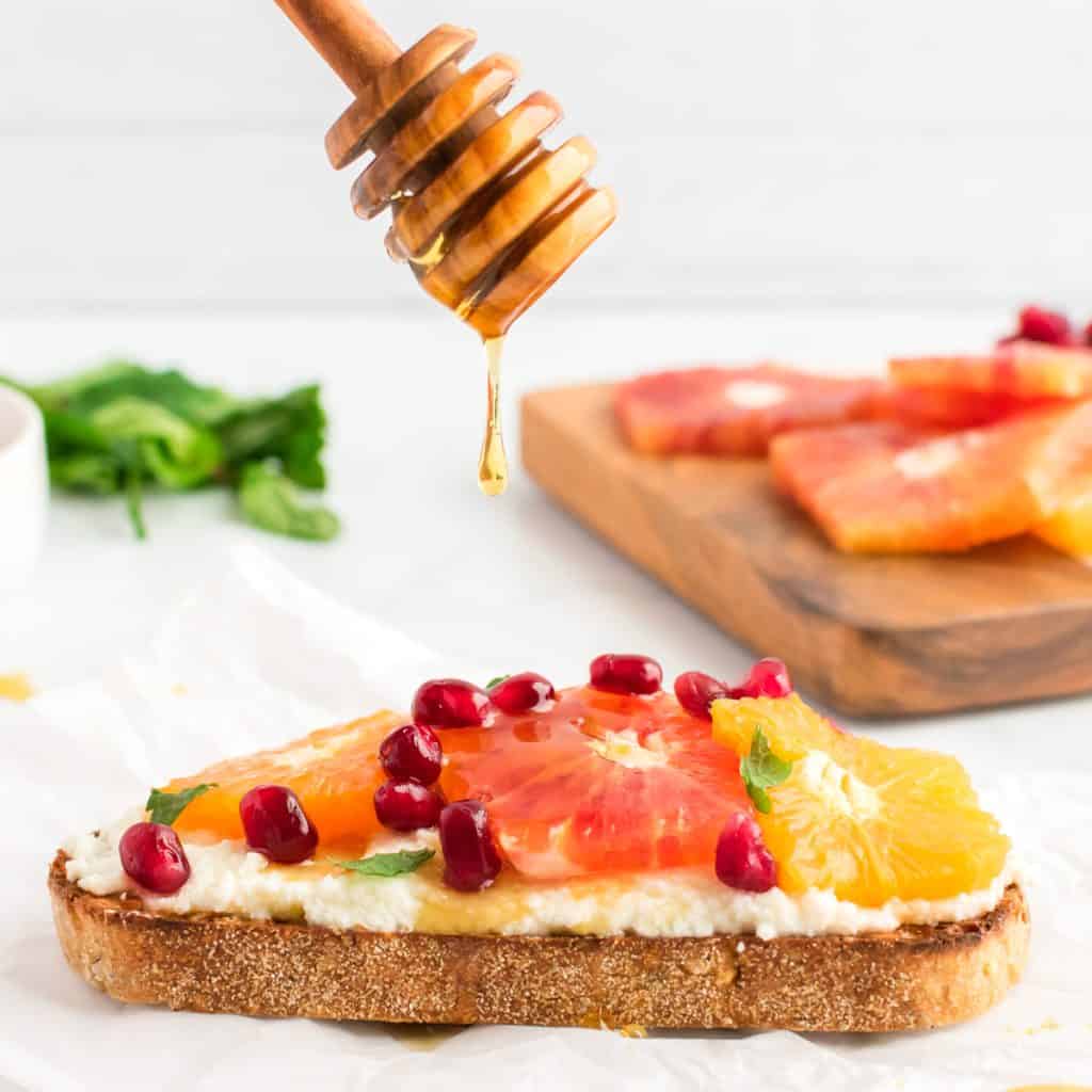 A slice of ricotta toast topped with citrus, with a honey spoon dripping a drip of honey onto it.