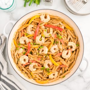 A white skillet on a marble background. The skillet contains shrimp rasta pasta, dotted with green onions. More green onions, salt and pepper, and a stack of plates and forks sit to the side.