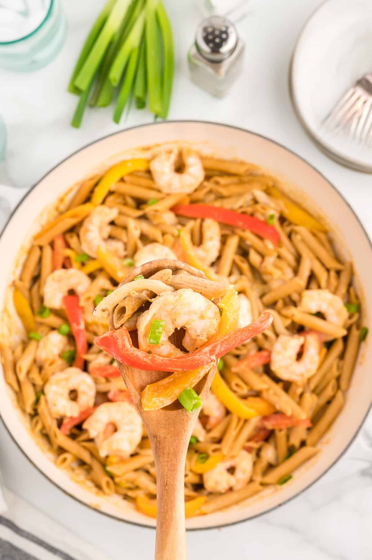 A wooden spoon holding a scoop of shrimp, peppers and penne pasta. A pan of shrimp rasta pasta is in the background.