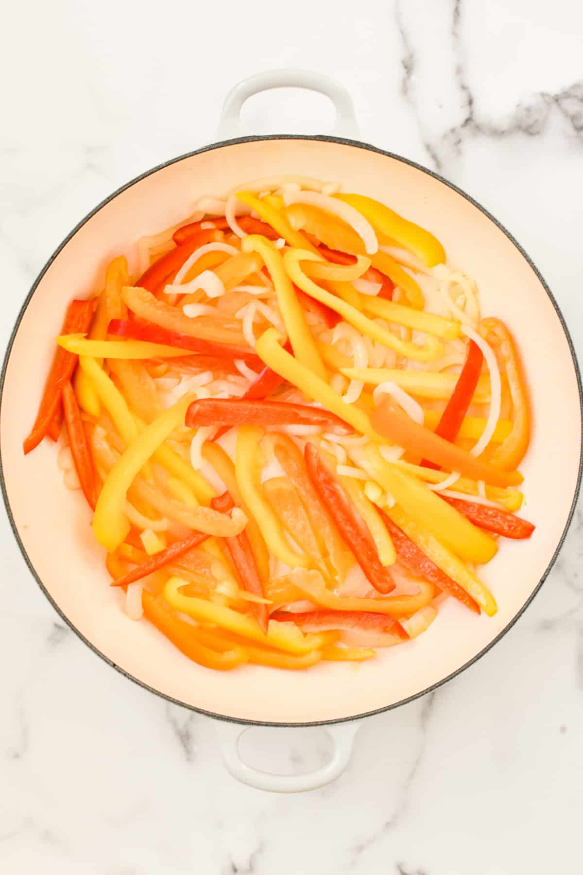 A white skillet on a marble background. The skillet contains raw onions and peppers.