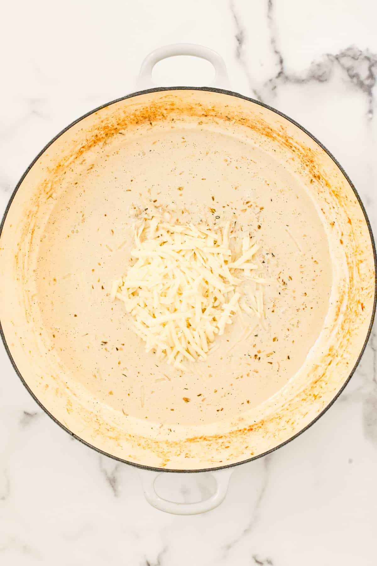 A white skillet on a marble background. The skillet contains cream with shredded cheese on top, not yet melted in.