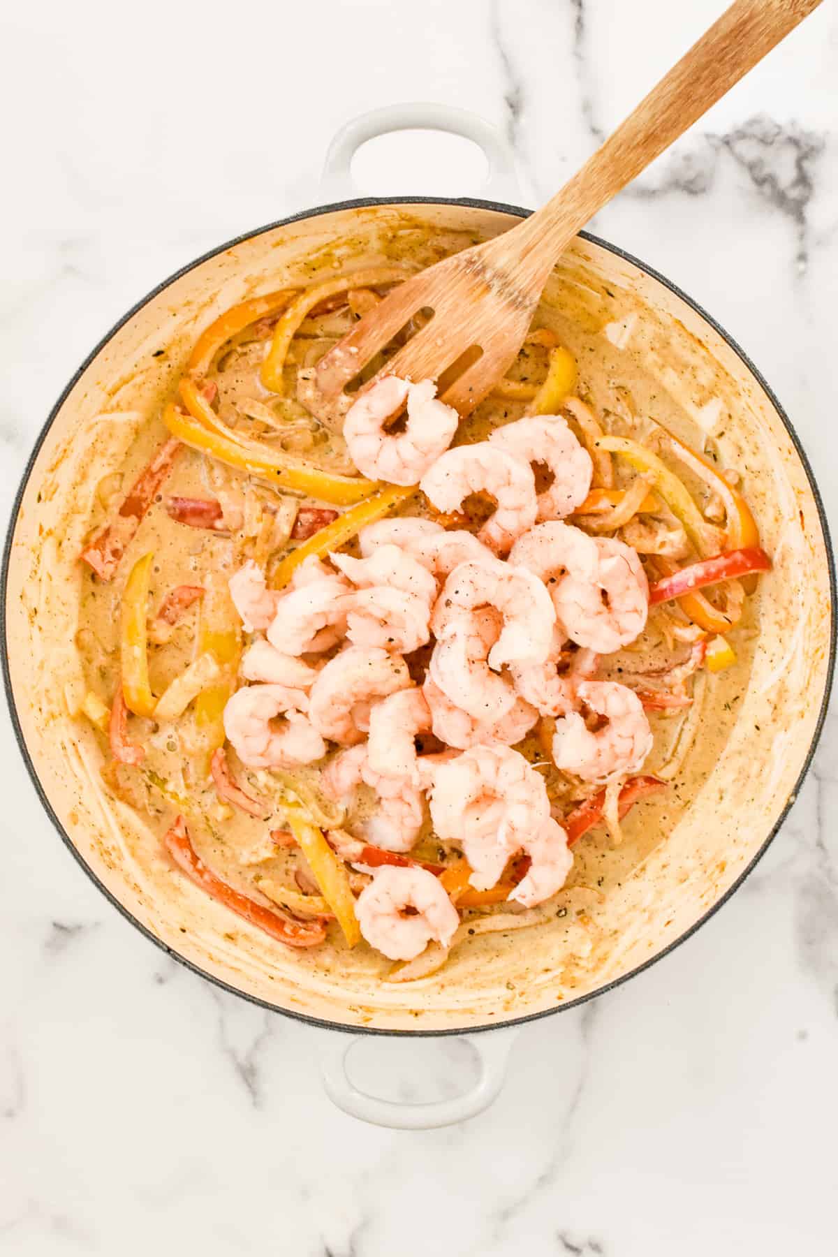 A white skillet on a marble background. The skillet contains jerk Alfredo sauce, peppers, onions and cooked shrimp.