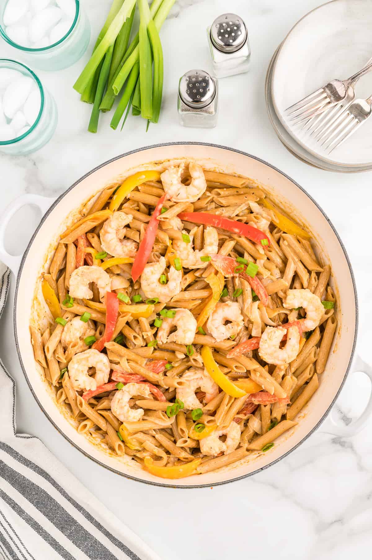 A white skillet on a marble background. The skillet contains shrimp rasta pasta, dotted with green onions. More green onions, salt and pepper, and a stack of plates and forks sit to the side.