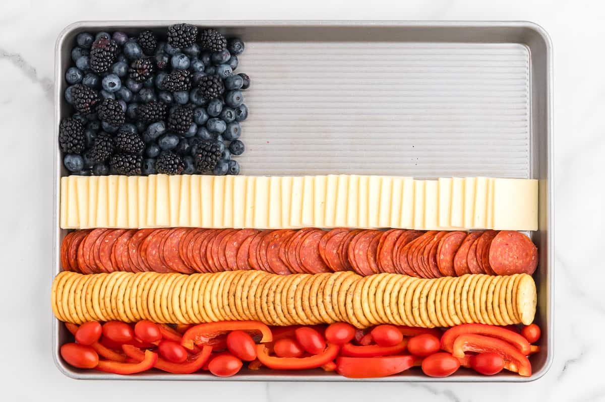 A rimmed sheet pan on a marble background. The sheet pan contains a charcuterie board flag, with 4 alternating red and white stripes and a blue square in the top left corner. The top right portion of the sheet pan is still empty.