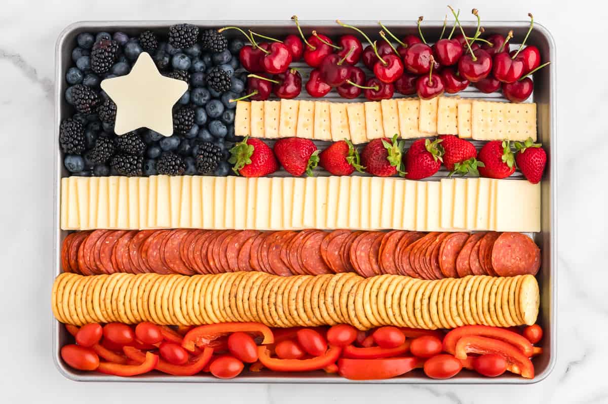 A sheet pan with a charcuterie board in the shape of the American flag. The flag has alternating red and white strips of cheese, crackers, pepperoni, fruits and vegetables. The top right is a blue section of blueberries and blackberries with a white cheese star.