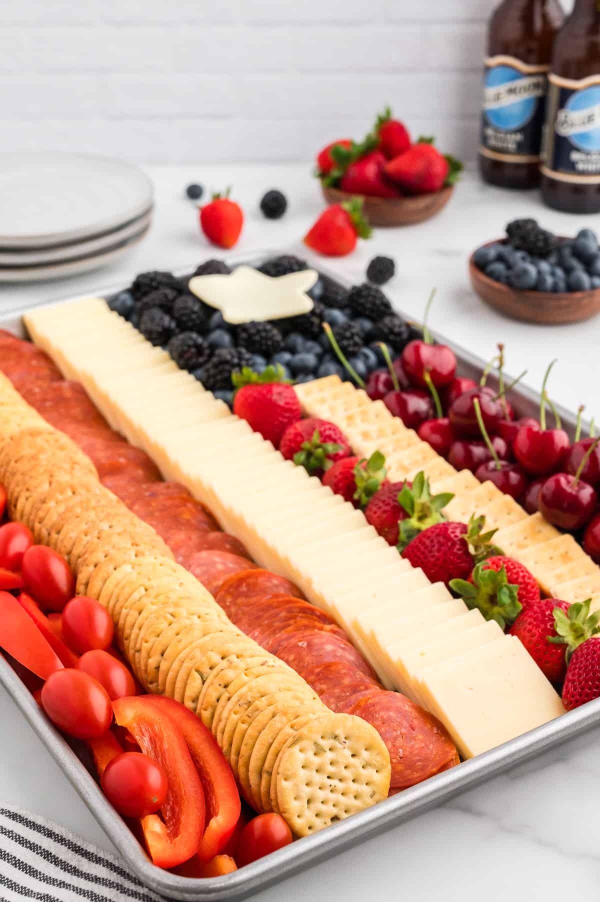 A side view of an American flag charcuterie board, with two beers, a stack of plates and bowls of fruit in the background.