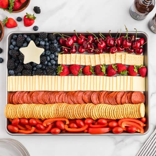 A sheet pan with a charcuterie board in the shape of the American flag. The flag has alternating red and white strips of cheese, crackers, pepperoni, fruits and vegetables. The top right is a blue section of blueberries and blackberries with a white cheese star.